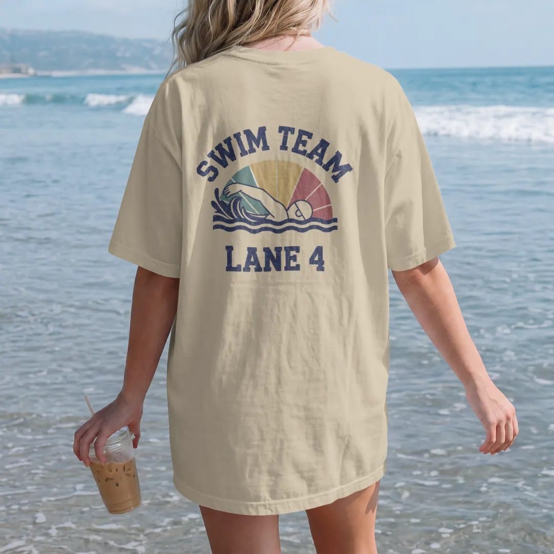 Woman on the beach in a Sandstone tee, back view: a swimmer mid freestyle stroke with stylized splashing waves and a lane rope, with "SWIM TEAM" and "LANE 4" in chunky faded collegiate block letters, printed in faded varsity navy, washed red,…