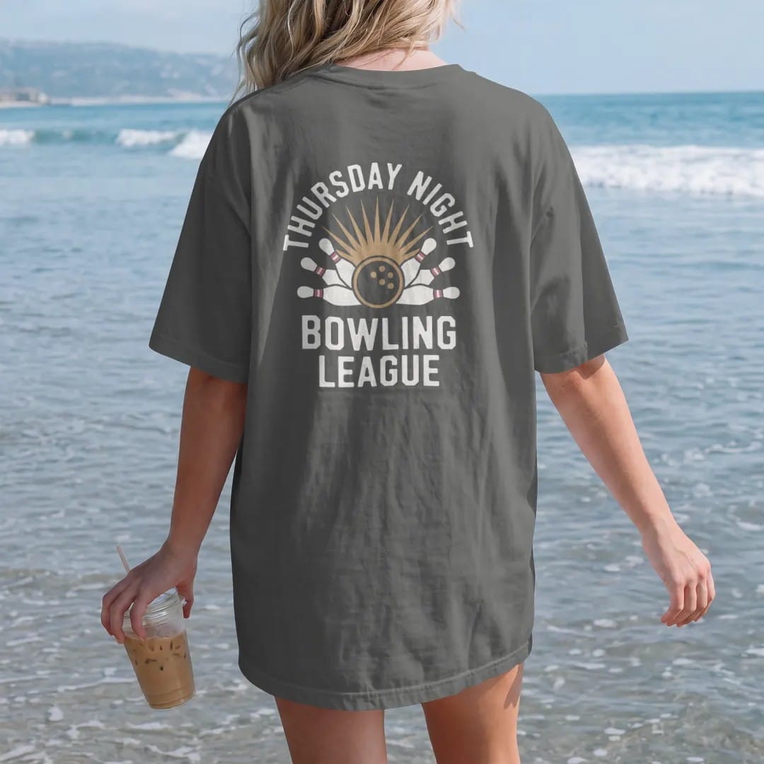 Woman on the beach in a Pepper tee, back view: a bowling ball crashing into scattering pins with a starburst, with "THURSDAY NIGHT" and "BOWLING LEAGUE" in chunky 1970s swash script, printed in faded bleacher cream, mustard gold, washed red and…