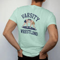 Man wearing a Chalky Mint tee, seen from the back: two wrestlers locked in a classic neutral stance inside a mat circle, with "VARSITY" and "WRESTLING" in chunky faded collegiate block letters, printed in faded varsity navy, washed red, mustard…