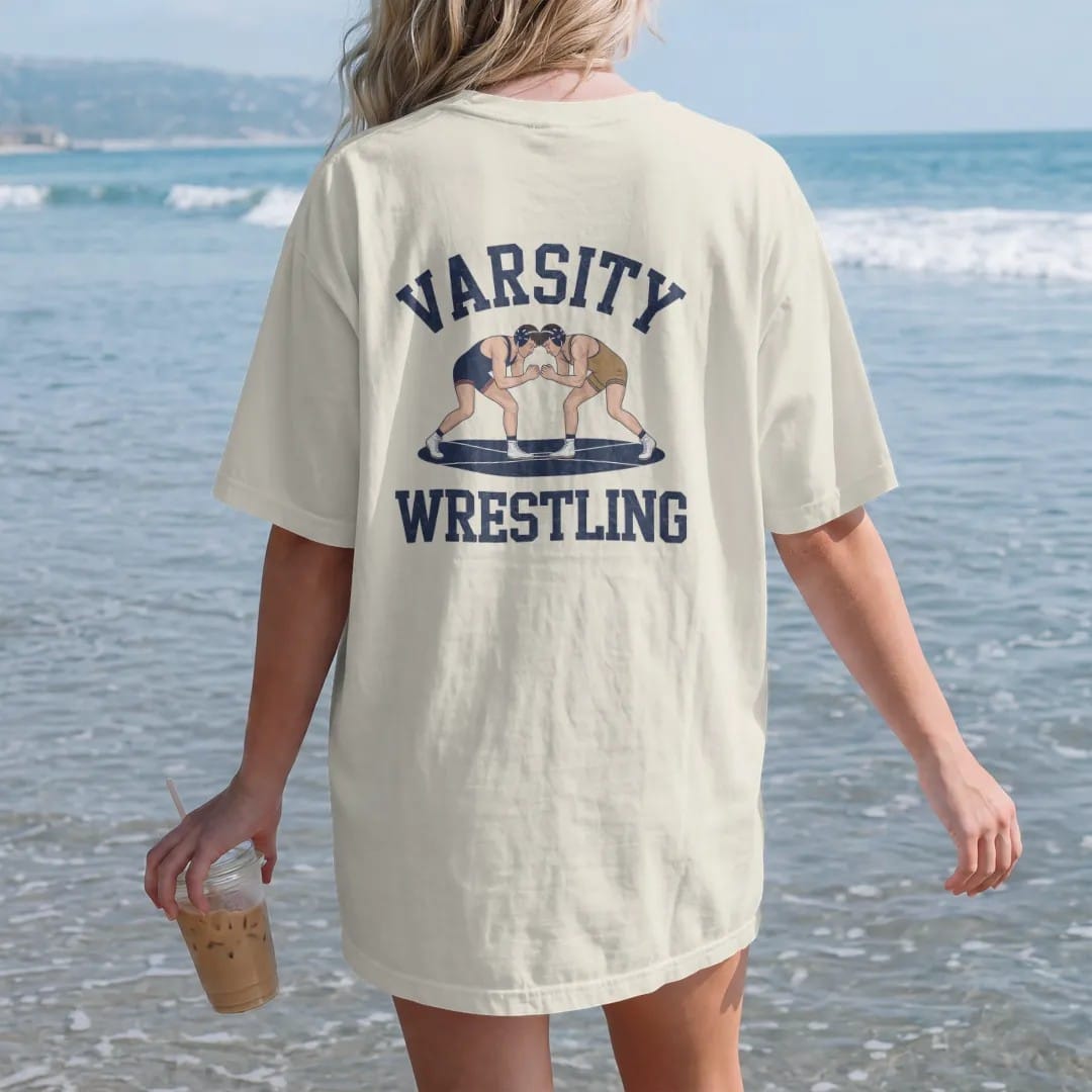 Woman on the beach in a Ivory tee, back view: two wrestlers locked in a classic neutral stance inside a mat circle, with "VARSITY" and "WRESTLING" in chunky faded collegiate block letters, printed in faded varsity navy, washed red, mustard gold and…