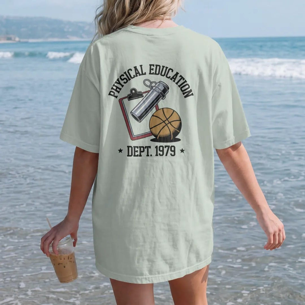 Woman on the beach in a Bay tee, back view: a chrome whistle on a lanyard over a clipboard and a rubber playground ball, with "PHYSICAL EDUCATION" and "DEPT. 1979" in chunky faded collegiate block letters, printed in faded varsity navy, washed red,…