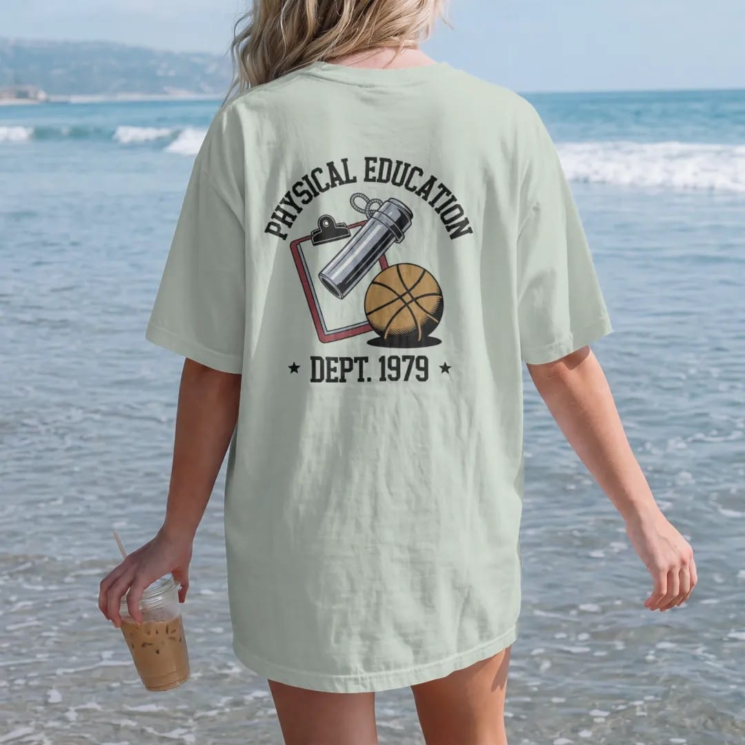 Woman on the beach in a Bay tee, back view: a chrome whistle on a lanyard over a clipboard and a rubber playground ball, with "PHYSICAL EDUCATION" and "DEPT. 1979" in chunky faded collegiate block letters, printed in faded varsity navy, washed red,…