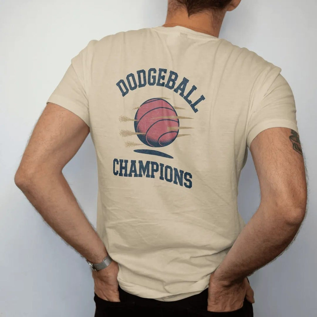 Man wearing a Sandstone tee, seen from the back: a red rubber playground ball flying with dust puffs and motion lines, with "DODGEBALL" and "CHAMPIONS" in chunky rounded 70s block letters, printed in faded varsity navy, washed red, mustard gold and…