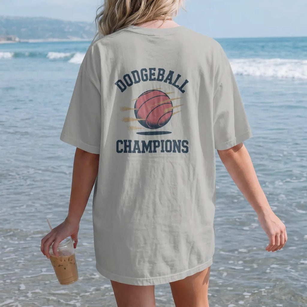 Woman on the beach in a Granite tee, back view: a red rubber playground ball flying with dust puffs and motion lines, with "DODGEBALL" and "CHAMPIONS" in chunky rounded 70s block letters, printed in faded varsity navy, washed red, mustard gold and…