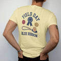 Man wearing a Butter tee, seen from the back: a blue ribbon rosette above a tug-of-war rope and an egg on a spoon, with "FIELD DAY" and "BLUE RIBBON" in chunky rounded 70s block letters, printed in faded varsity navy, washed red, mustard gold and…