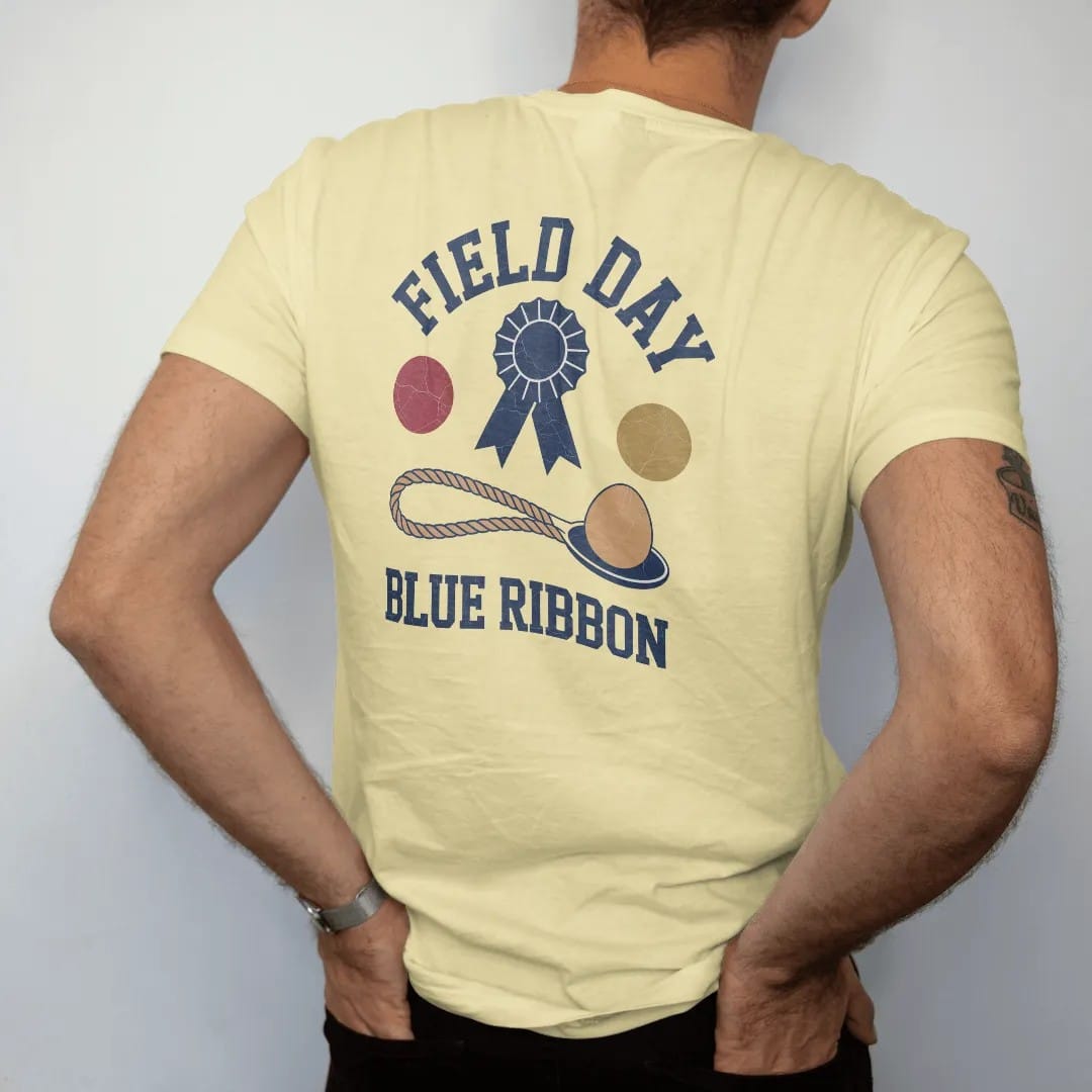 Man wearing a Butter tee, seen from the back: a blue ribbon rosette above a tug-of-war rope and an egg on a spoon, with "FIELD DAY" and "BLUE RIBBON" in chunky rounded 70s block letters, printed in faded varsity navy, washed red, mustard gold and…