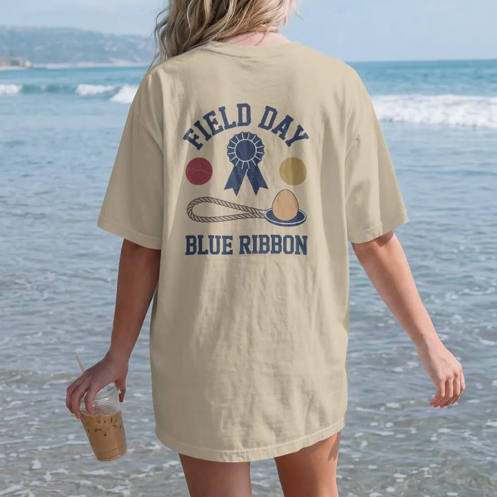 Woman on the beach in a Sandstone tee, back view: a blue ribbon rosette above a tug-of-war rope and an egg on a spoon, with "FIELD DAY" and "BLUE RIBBON" in chunky rounded 70s block letters, printed in faded varsity navy, washed red, mustard gold…