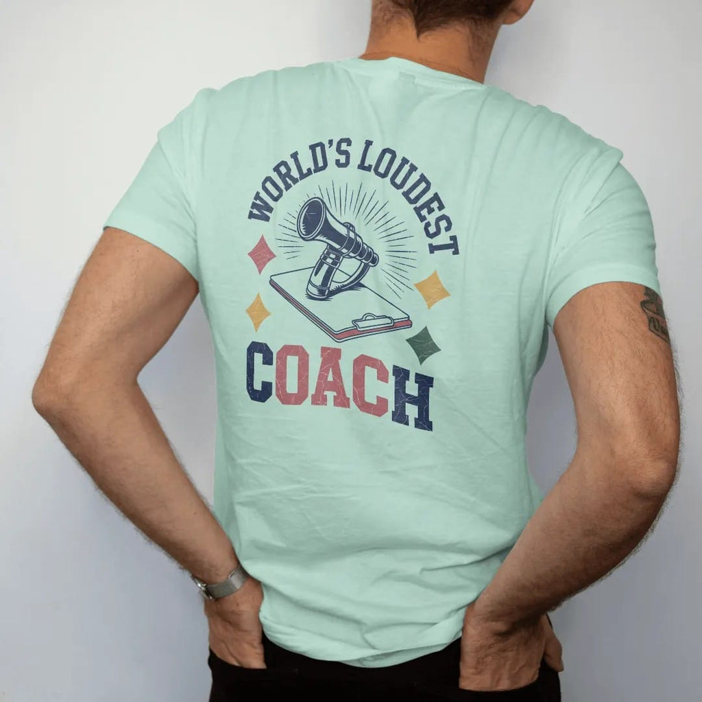 Man wearing a Chalky Mint tee, seen from the back: a chrome whistle with sound-wave lines bursting out above a clipboard, with "WORLD'S LOUDEST" and "COACH" in chunky faded collegiate block letters, printed in faded varsity navy, washed red,…