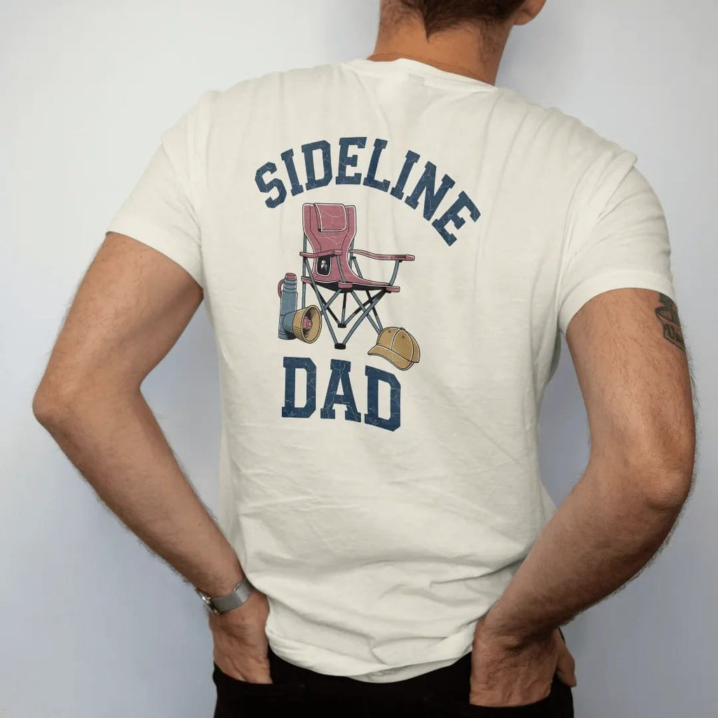 Man wearing a Ivory tee, seen from the back: a folding stadium chair with a thermos, a megaphone and a team cap, with "SIDELINE" and "DAD" in chunky 1970s swash script, printed in faded varsity navy, washed red, mustard gold and pine green with a…