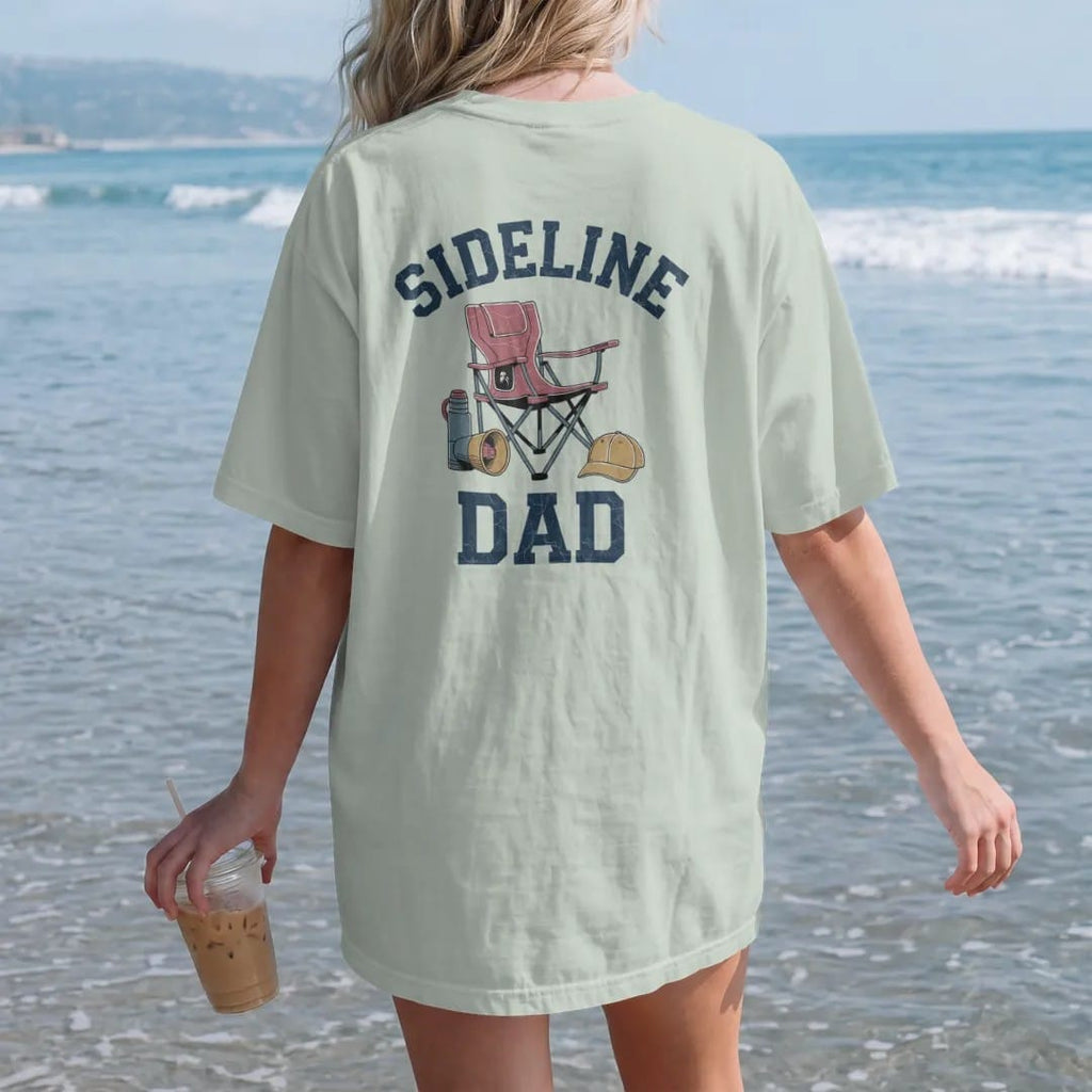 Woman on the beach in a Bay tee, back view: a folding stadium chair with a thermos, a megaphone and a team cap, with "SIDELINE" and "DAD" in chunky 1970s swash script, printed in faded varsity navy, washed red, mustard gold and pine green with a…