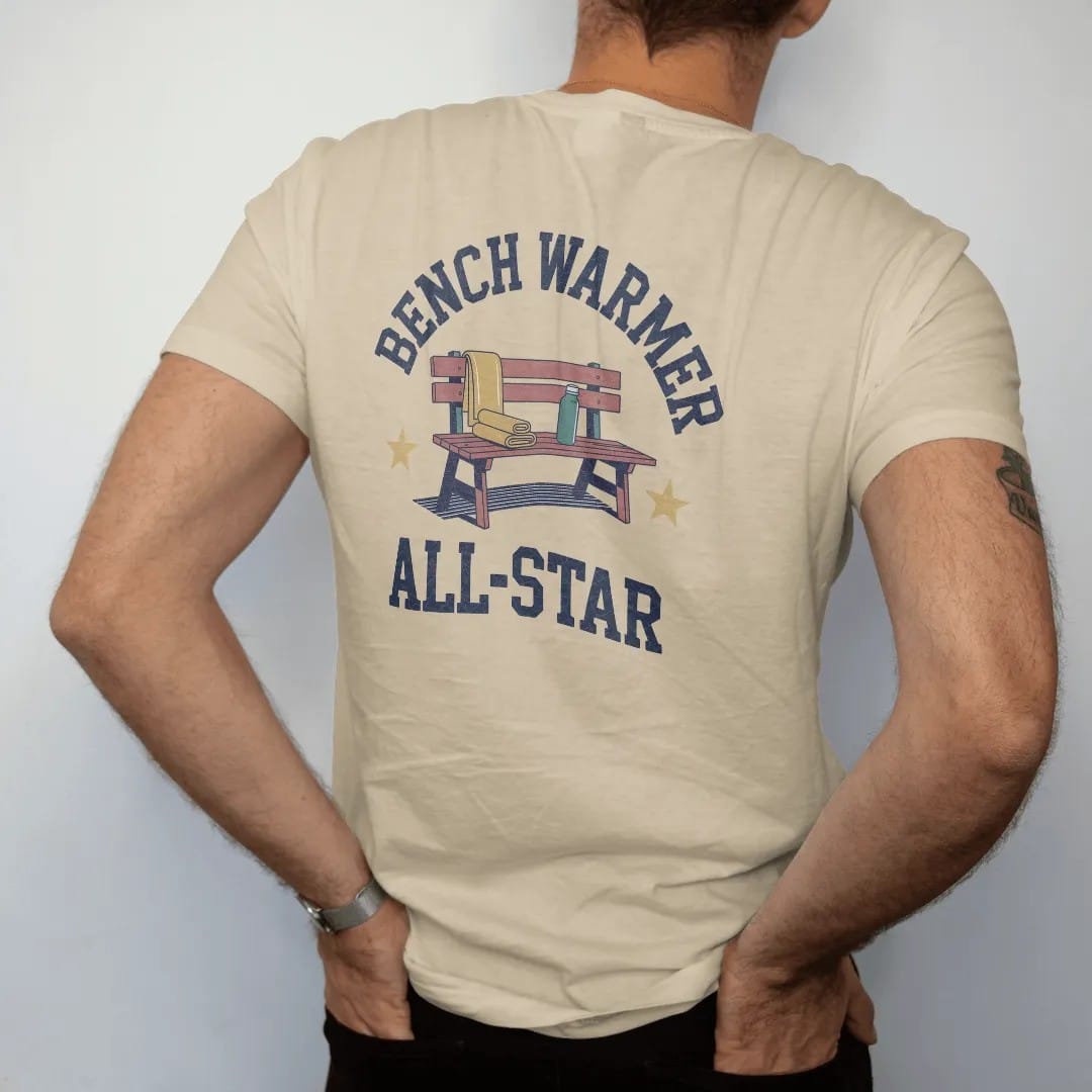 Man wearing a Sandstone tee, seen from the back: a wooden team bench with a folded towel, a water bottle and a gold star, with "BENCH WARMER" and "ALL-STAR" in chunky rounded 70s block letters, printed in faded varsity navy, washed red, mustard…