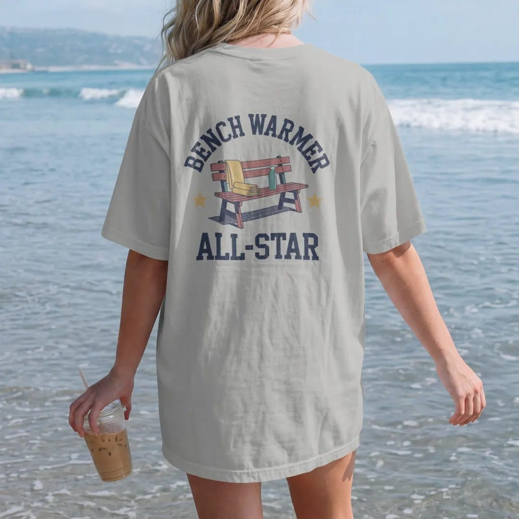 Woman on the beach in a Granite tee, back view: a wooden team bench with a folded towel, a water bottle and a gold star, with "BENCH WARMER" and "ALL-STAR" in chunky rounded 70s block letters, printed in faded varsity navy, washed red, mustard gold…