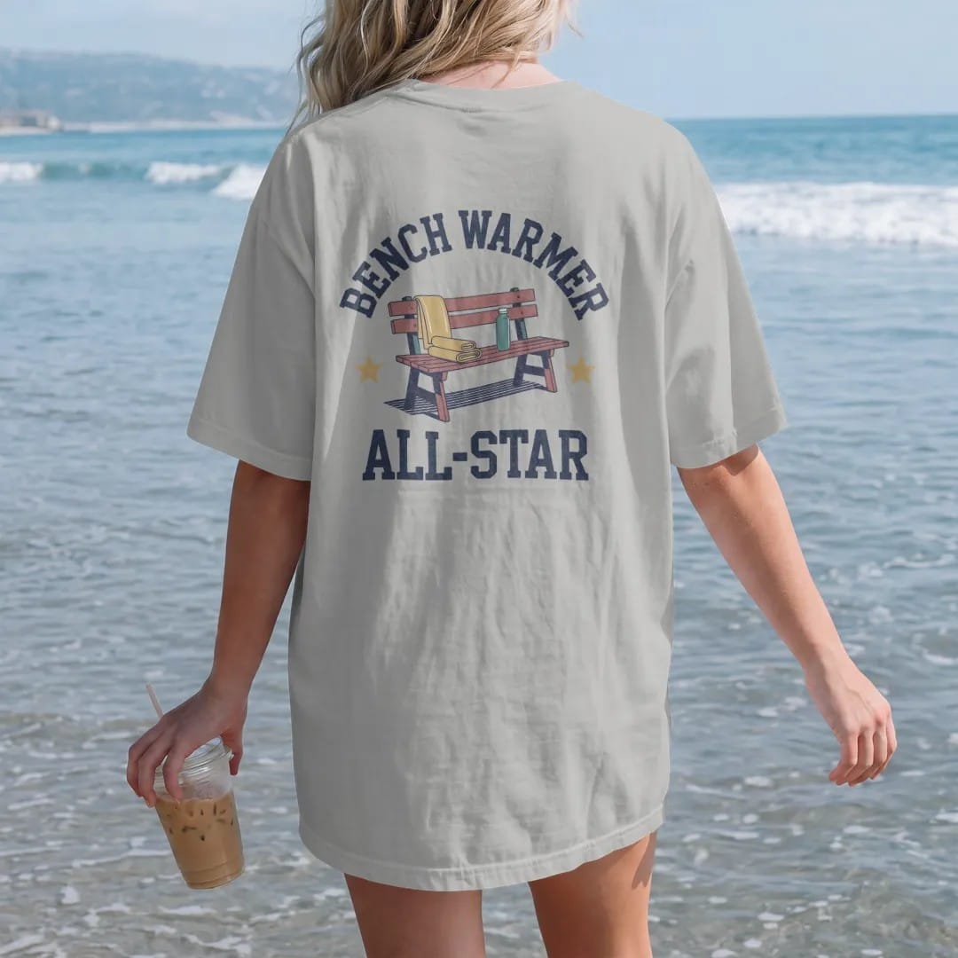 Woman on the beach in a Granite tee, back view: a wooden team bench with a folded towel, a water bottle and a gold star, with "BENCH WARMER" and "ALL-STAR" in chunky rounded 70s block letters, printed in faded varsity navy, washed red, mustard gold…