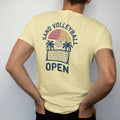 Man wearing a Butter tee, seen from the back: a volleyball being spiked over a net with a sun and palm trees, with "SAND VOLLEYBALL" and "OPEN" in chunky 1970s swash script, printed in faded varsity navy, washed red, mustard gold and pine green…