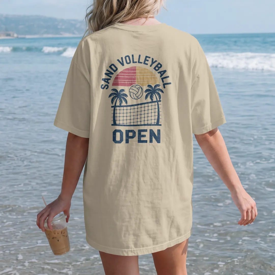 Woman on the beach in a Sandstone tee, back view: a volleyball being spiked over a net with a sun and palm trees, with "SAND VOLLEYBALL" and "OPEN" in chunky 1970s swash script, printed in faded varsity navy, washed red, mustard gold and pine green…