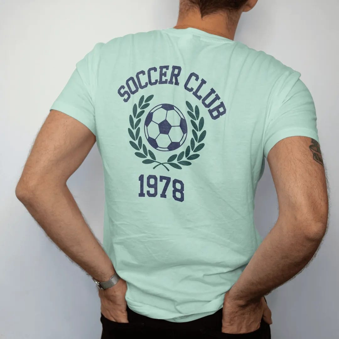 Man wearing a Chalky Mint tee, seen from the back: a classic paneled soccer ball with motion lines inside a laurel wreath, with "SOCCER CLUB" and "1978" in chunky faded collegiate block letters, printed in faded varsity navy, washed red, mustard…
