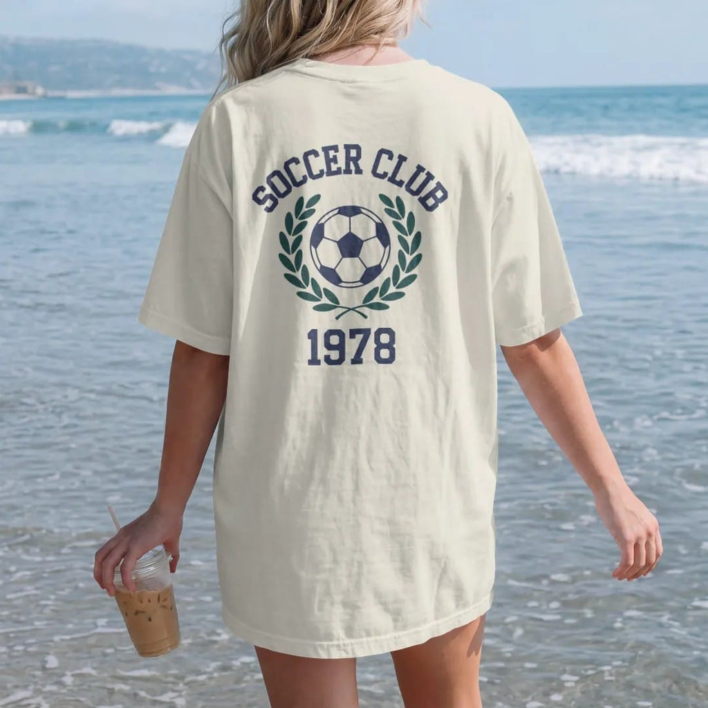 Woman on the beach in a Ivory tee, back view: a classic paneled soccer ball with motion lines inside a laurel wreath, with "SOCCER CLUB" and "1978" in chunky faded collegiate block letters, printed in faded varsity navy, washed red, mustard gold…