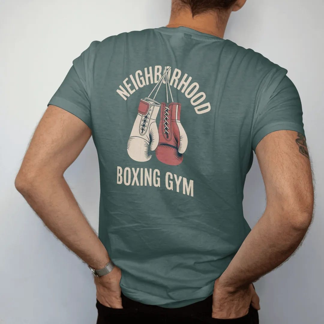 Man wearing a Blue Spruce tee, seen from the back: a pair of laced vintage boxing gloves hanging by their laces, with "NEIGHBORHOOD" and "BOXING GYM" in faded condensed 70s fight-poster letters, printed in faded bleacher cream, mustard gold, washed…