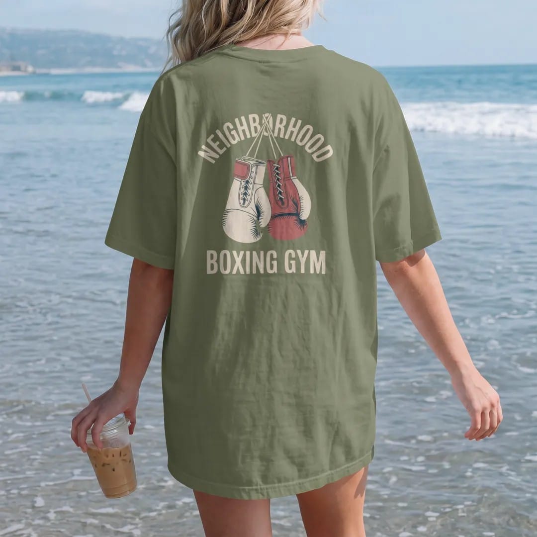 Woman on the beach in a Moss tee, back view: a pair of laced vintage boxing gloves hanging by their laces, with "NEIGHBORHOOD" and "BOXING GYM" in faded condensed 70s fight-poster letters, printed in faded bleacher cream, mustard gold, washed red…