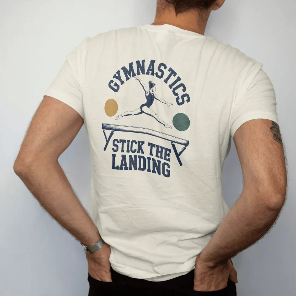 Man wearing a Ivory tee, seen from the back: a gymnast mid leap on a balance beam with motion arcs, with "GYMNASTICS" and "STICK THE LANDING" in chunky 1970s swash script, printed in faded varsity navy, washed red, mustard gold and pine green with…