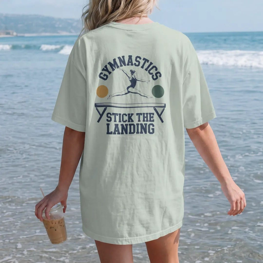 Woman on the beach in a Bay tee, back view: a gymnast mid leap on a balance beam with motion arcs, with "GYMNASTICS" and "STICK THE LANDING" in chunky 1970s swash script, printed in faded varsity navy, washed red, mustard gold and pine green with a…