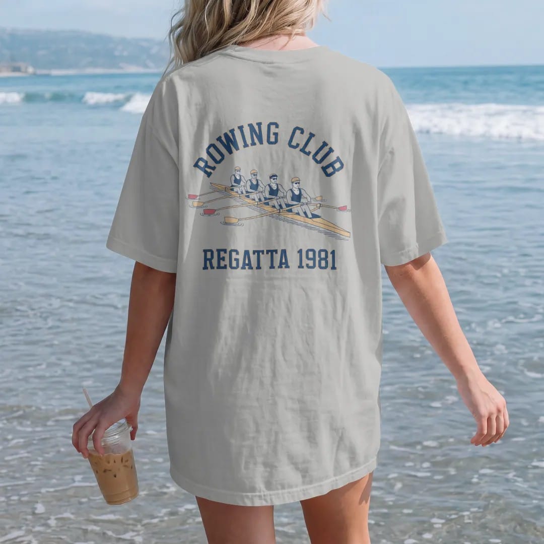 Woman on the beach in a Granite tee, back view: a long rowing shell with four rowers pulling in sync on rippled water, with "ROWING CLUB" and "REGATTA 1981" in chunky faded collegiate block letters, printed in faded varsity navy, washed red,…