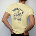 Man wearing a Butter tee, seen from the back: a vintage road bicycle with drop handlebars and a cycling cap hanging off it, with "VELO CLUB" and "EST. 1976" in chunky 1970s swash script, printed in faded varsity navy, washed red, mustard gold and…