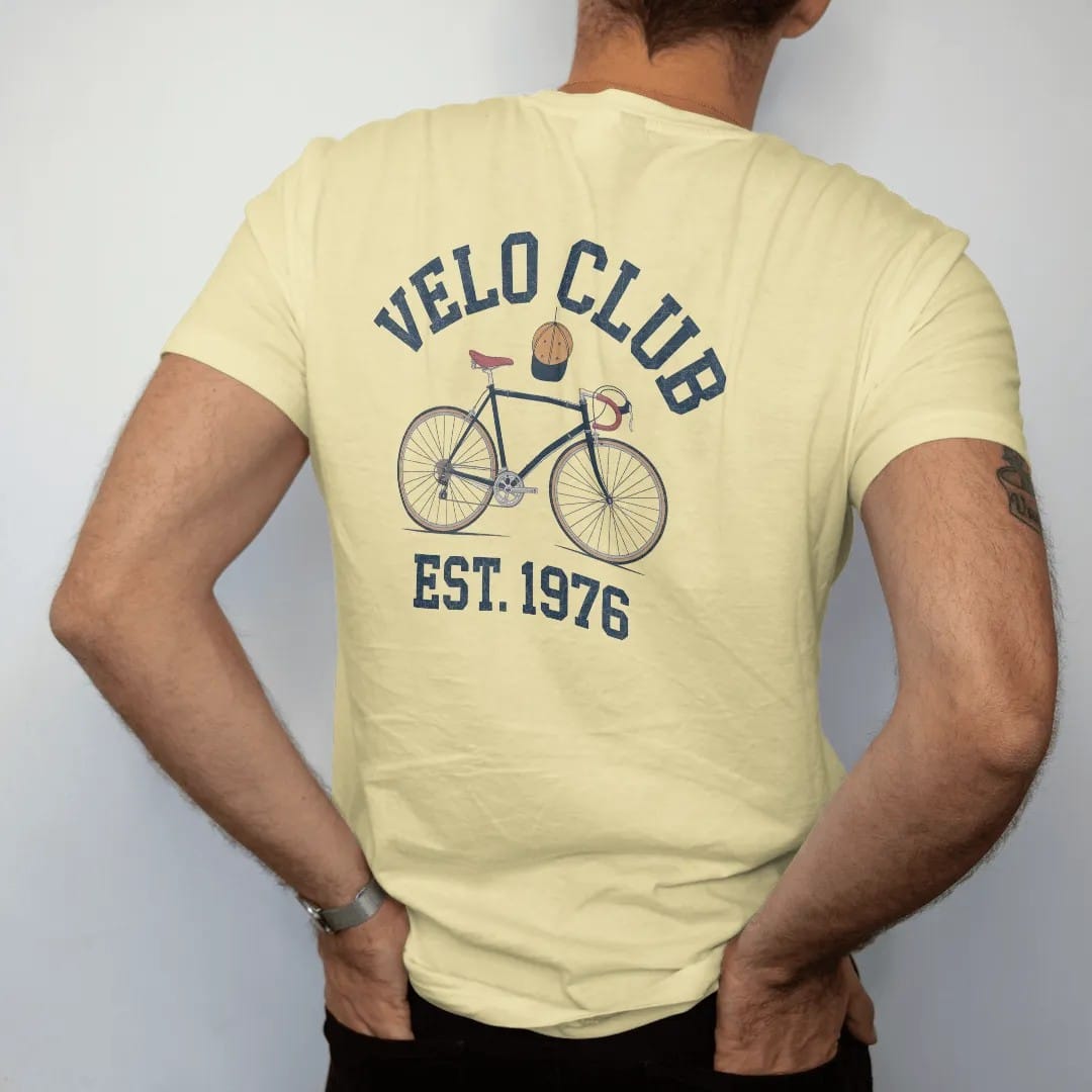 Man wearing a Butter tee, seen from the back: a vintage road bicycle with drop handlebars and a cycling cap hanging off it, with "VELO CLUB" and "EST. 1976" in chunky 1970s swash script, printed in faded varsity navy, washed red, mustard gold and…