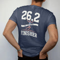 Man wearing a Navy tee, seen from the back: a runner breaking the finish-line tape with arms raised, with "26.2" and "FINISHER" in chunky faded collegiate block letters, printed in faded bleacher cream, mustard gold, washed red and dusty sky blue…