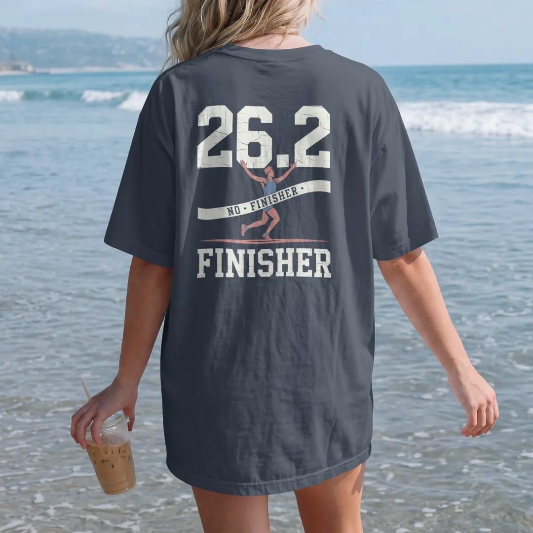 Woman on the beach in a Midnight tee, back view: a runner breaking the finish-line tape with arms raised, with "26.2" and "FINISHER" in chunky faded collegiate block letters, printed in faded bleacher cream, mustard gold, washed red and dusty sky…
