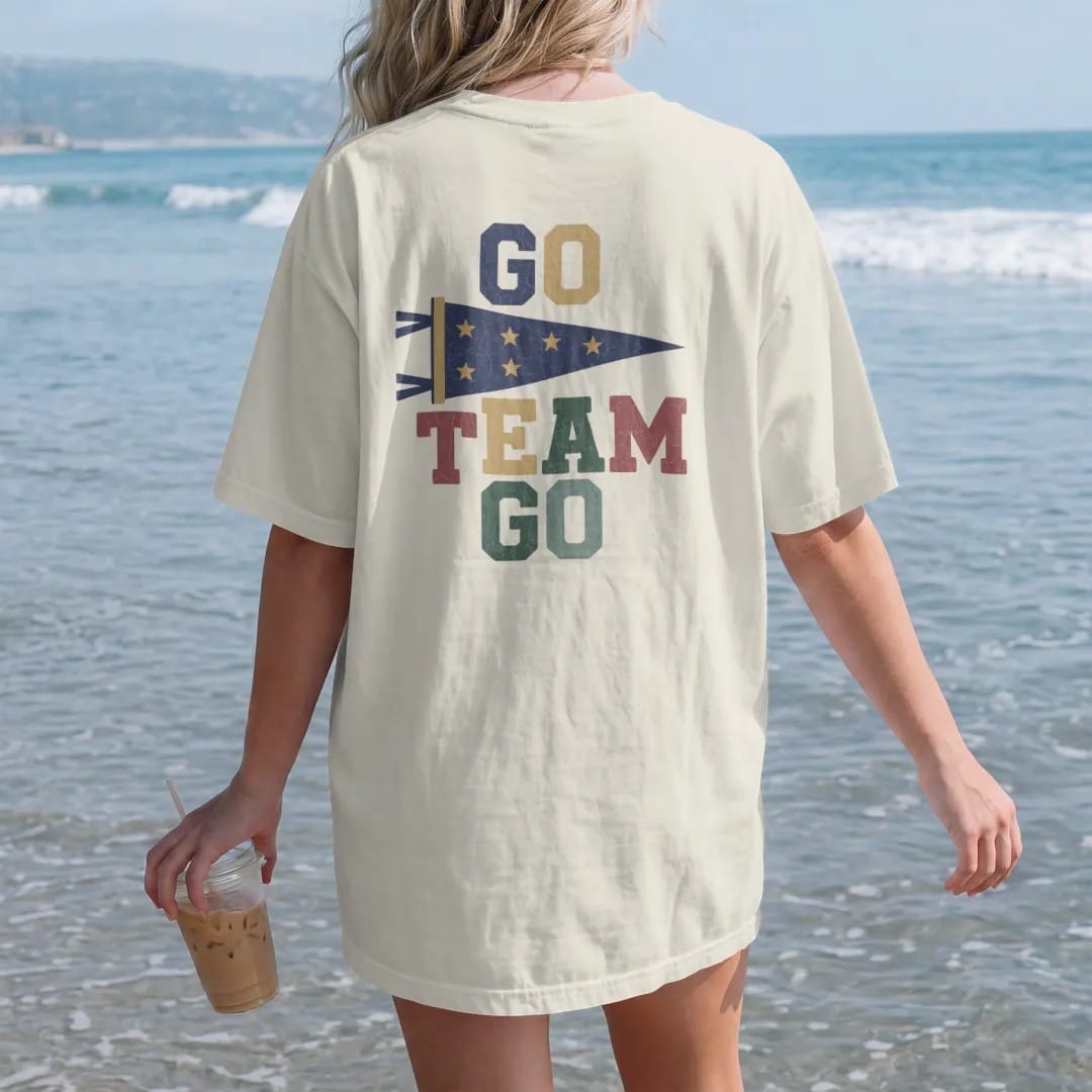 Woman on the beach in a Ivory tee, back view: a classic felt pennant waving on a stick with stars, with "GO TEAM GO" in chunky rounded 70s block letters, printed in faded varsity navy, washed red, mustard gold and pine green with a cracked, worn…