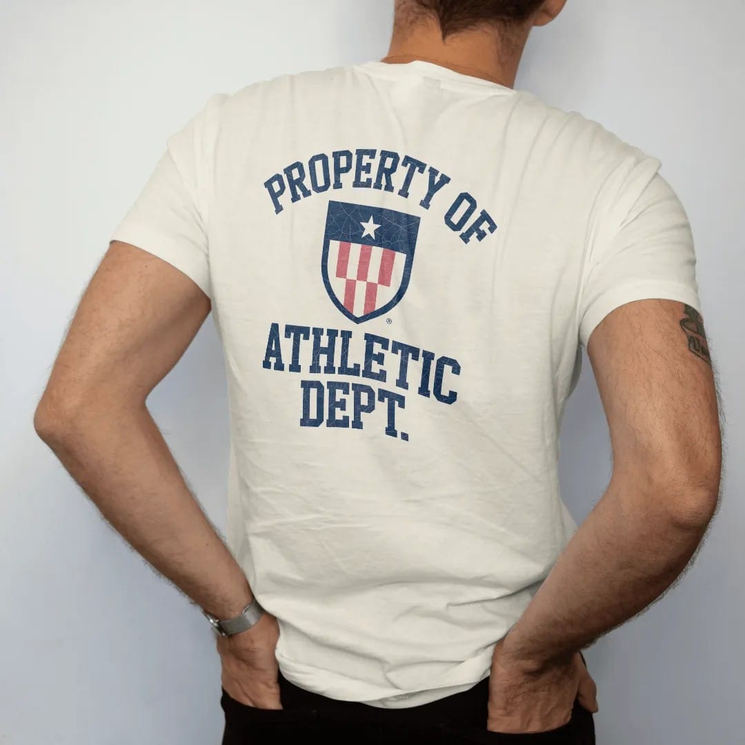 Man wearing a Ivory tee, seen from the back: a simple athletic shield emblem with a star, with "PROPERTY OF" and "ATHLETIC DEPT." in chunky faded collegiate block letters, printed in faded varsity navy, washed red, mustard gold and pine green with…
