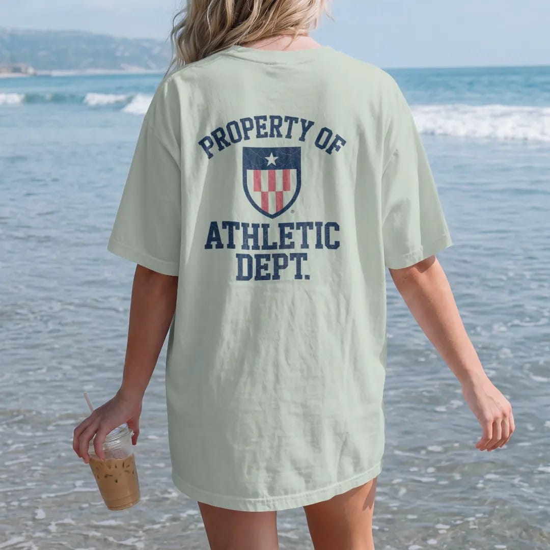 Woman on the beach in a Bay tee, back view: a simple athletic shield emblem with a star, with "PROPERTY OF" and "ATHLETIC DEPT." in chunky faded collegiate block letters, printed in faded varsity navy, washed red, mustard gold and pine green with a…