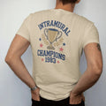 Man wearing a Sandstone tee, seen from the back: a gold trophy cup with laurel leaves and stars, with "INTRAMURAL" and "CHAMPIONS" and "1983" in chunky faded collegiate block letters, printed in faded varsity navy, washed red, mustard gold and pine…