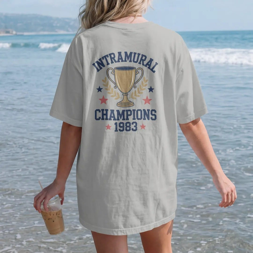 Woman on the beach in a Granite tee, back view: a gold trophy cup with laurel leaves and stars, with "INTRAMURAL" and "CHAMPIONS" and "1983" in chunky faded collegiate block letters, printed in faded varsity navy, washed red, mustard gold and pine…