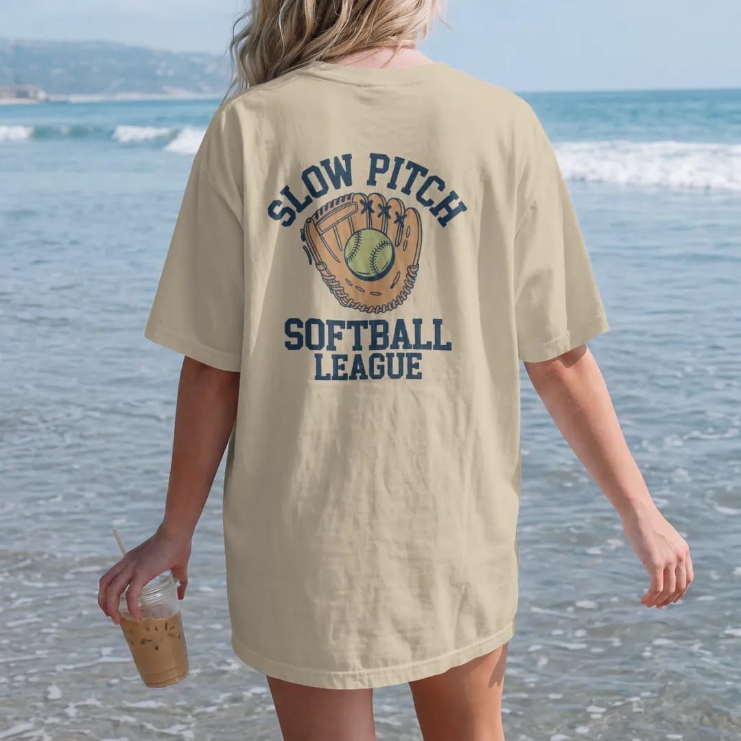 Woman on the beach in a Sandstone tee, back view: a softball and leather glove with dirt puffs, with "SLOW PITCH" and "SOFTBALL LEAGUE" in chunky 1970s swash script, printed in faded varsity navy, washed red, mustard gold and pine green with a…