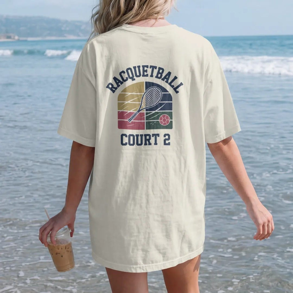 Woman on the beach in a Ivory tee, back view: a racquetball racquet and ball bouncing off a court wall with motion lines, with "RACQUETBALL" and "COURT 2" in chunky rounded 70s block letters, printed in faded varsity navy, washed red, mustard gold…