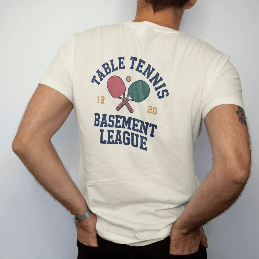 Man wearing a Ivory tee, seen from the back: two crossed paddles with a small ball bouncing over a table net, with "TABLE TENNIS" and "BASEMENT LEAGUE" in chunky faded collegiate block letters, printed in faded varsity navy, washed red, mustard…