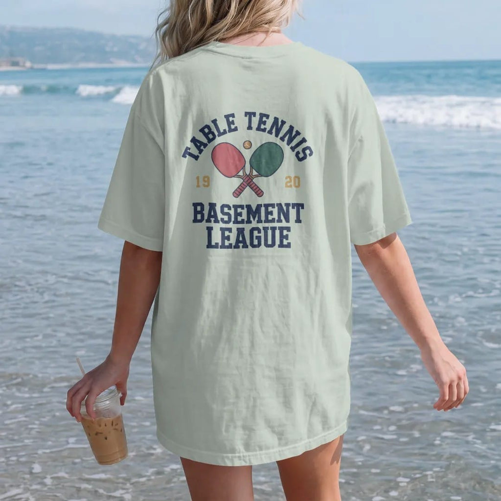 Woman on the beach in a Bay tee, back view: two crossed paddles with a small ball bouncing over a table net, with "TABLE TENNIS" and "BASEMENT LEAGUE" in chunky faded collegiate block letters, printed in faded varsity navy, washed red, mustard gold…