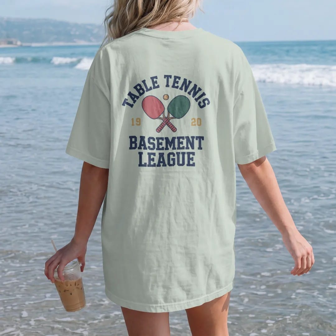 Woman on the beach in a Bay tee, back view: two crossed paddles with a small ball bouncing over a table net, with "TABLE TENNIS" and "BASEMENT LEAGUE" in chunky faded collegiate block letters, printed in faded varsity navy, washed red, mustard gold…