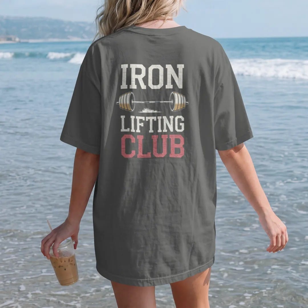 Woman on the beach in a Pepper tee, back view: a vintage barbell with round plates and a puff of chalk, with "IRON" and "LIFTING CLUB" in faded condensed 70s fight-poster letters, printed in faded bleacher cream, mustard gold, washed red and dusty…