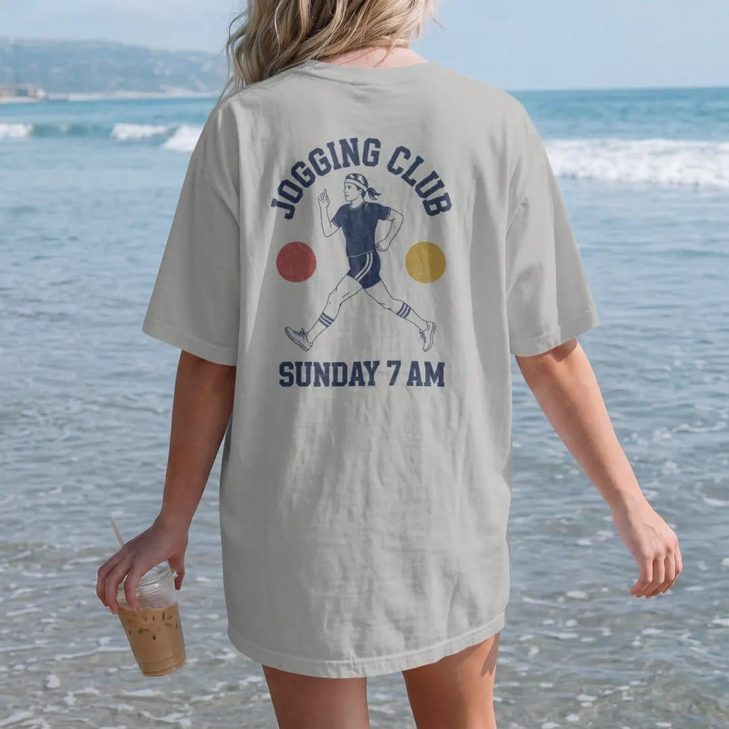 Woman on the beach in a Granite tee, back view: a 1970s jogger in a headband, short shorts and striped tube socks, with "JOGGING CLUB" and "SUNDAY 7 AM" in chunky 1970s swash script, printed in faded varsity navy, washed red, mustard gold and pine…