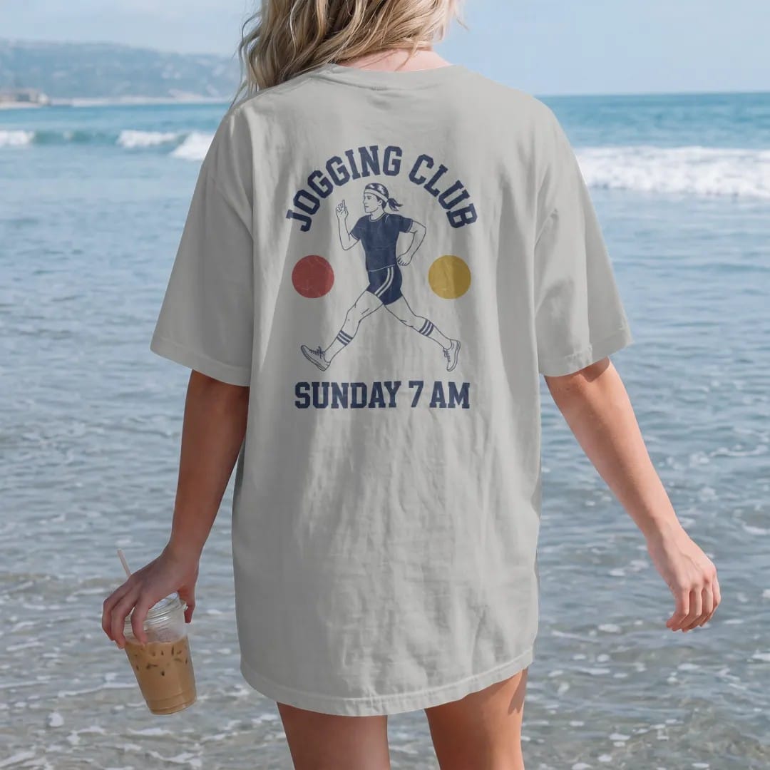 Woman on the beach in a Granite tee, back view: a 1970s jogger in a headband, short shorts and striped tube socks, with "JOGGING CLUB" and "SUNDAY 7 AM" in chunky 1970s swash script, printed in faded varsity navy, washed red, mustard gold and pine…
