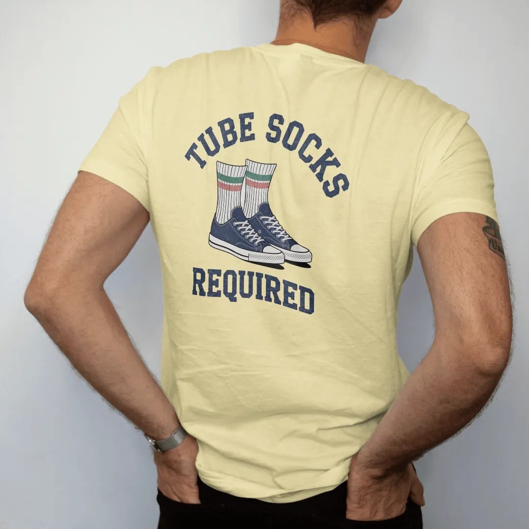 Man wearing a Butter tee, seen from the back: a pair of striped tube socks pulled high over sneakers, with "TUBE SOCKS" and "REQUIRED" in chunky rounded 70s block letters, printed in faded varsity navy, washed red, mustard gold and pine green with…