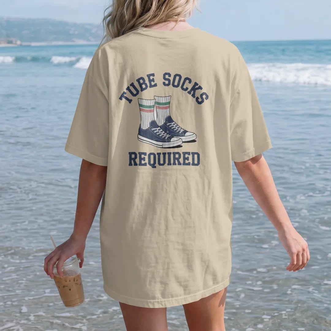 Woman on the beach in a Sandstone tee, back view: a pair of striped tube socks pulled high over sneakers, with "TUBE SOCKS" and "REQUIRED" in chunky rounded 70s block letters, printed in faded varsity navy, washed red, mustard gold and pine green…