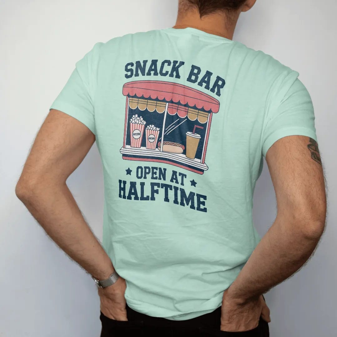 Man wearing a Chalky Mint tee, seen from the back: a vintage concession window with popcorn, a hot dog and a paper cup soda, with "SNACK BAR" and "OPEN AT HALFTIME" in chunky rounded 70s sign letters, printed in faded varsity navy, washed red,…