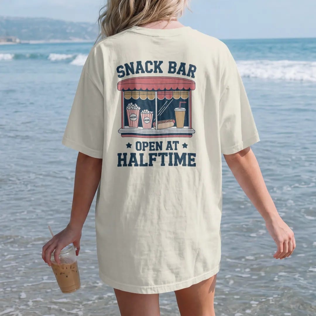 Woman on the beach in a Ivory tee, back view: a vintage concession window with popcorn, a hot dog and a paper cup soda, with "SNACK BAR" and "OPEN AT HALFTIME" in chunky rounded 70s sign letters, printed in faded varsity navy, washed red, mustard…