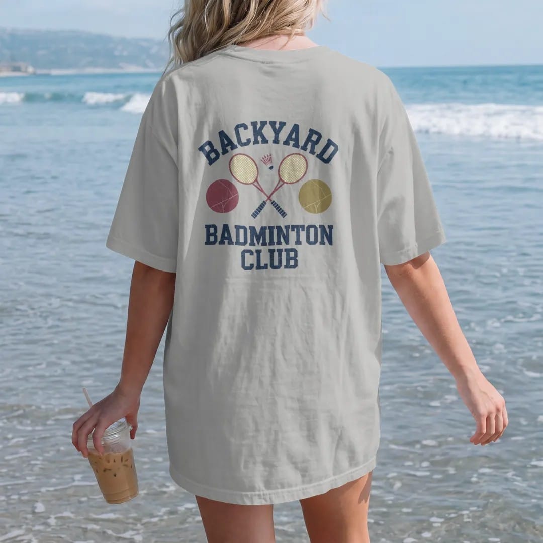 Woman on the beach in a Granite tee, back view: a shuttlecock and two racquets crossed over a backyard net, with "BACKYARD" and "BADMINTON CLUB" in chunky rounded 70s block letters, printed in faded varsity navy, washed red, mustard gold and pine…