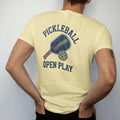 Man wearing a Butter tee, seen from the back: a paddle and a holed plastic ball with speed lines, with "PICKLEBALL" and "OPEN PLAY" in chunky faded collegiate block letters, printed in faded varsity navy, washed red, mustard gold and pine green…