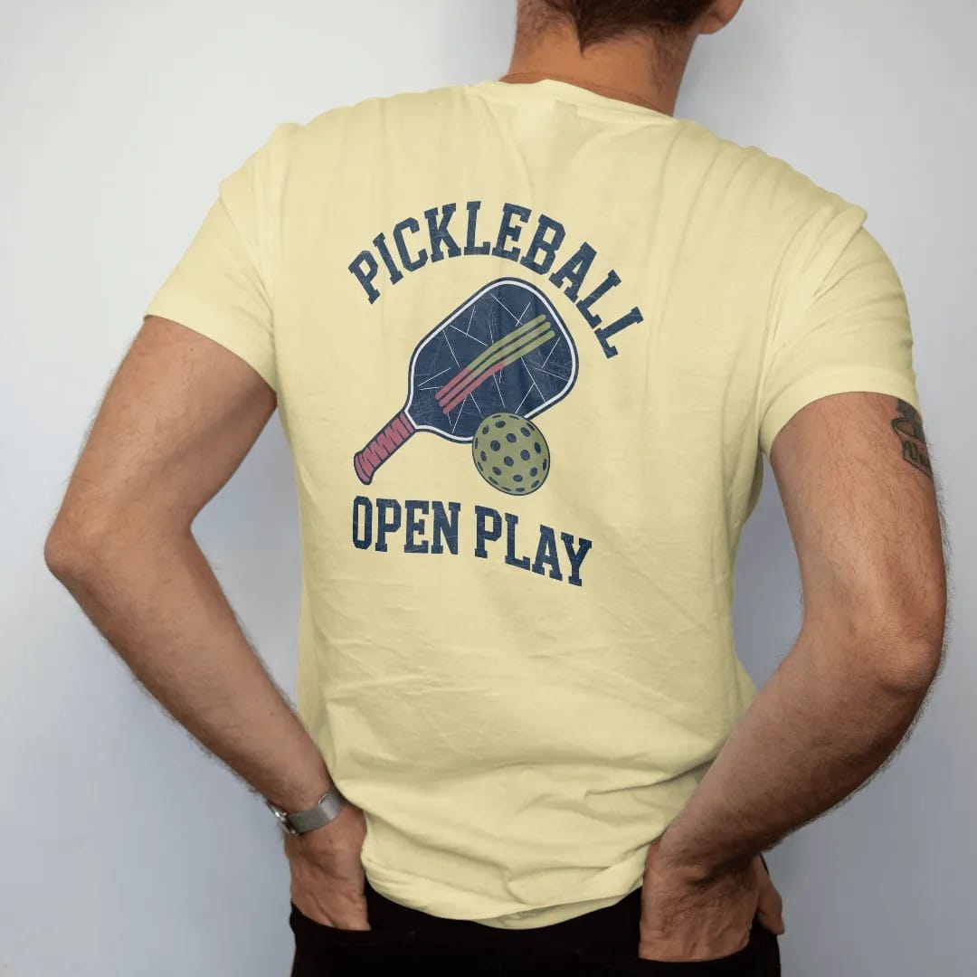 Man wearing a Butter tee, seen from the back: a paddle and a holed plastic ball with speed lines, with "PICKLEBALL" and "OPEN PLAY" in chunky faded collegiate block letters, printed in faded varsity navy, washed red, mustard gold and pine green…